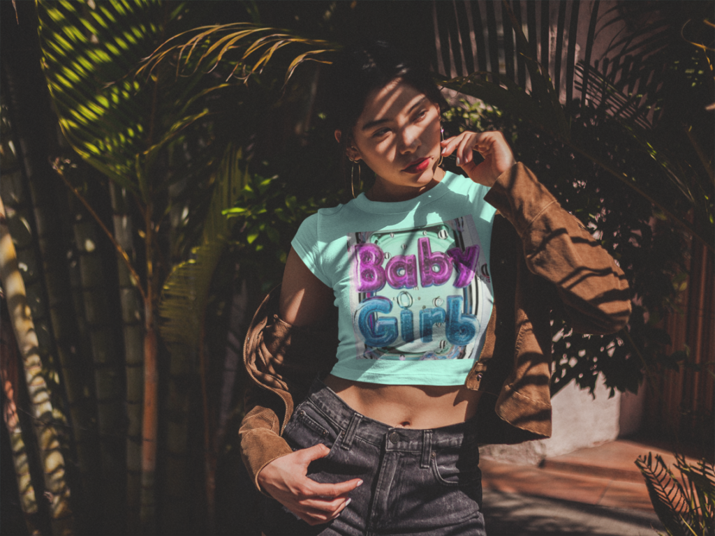 Girl wearing a crop-top t-shirt saying-Baby-girl under a palm tree.
